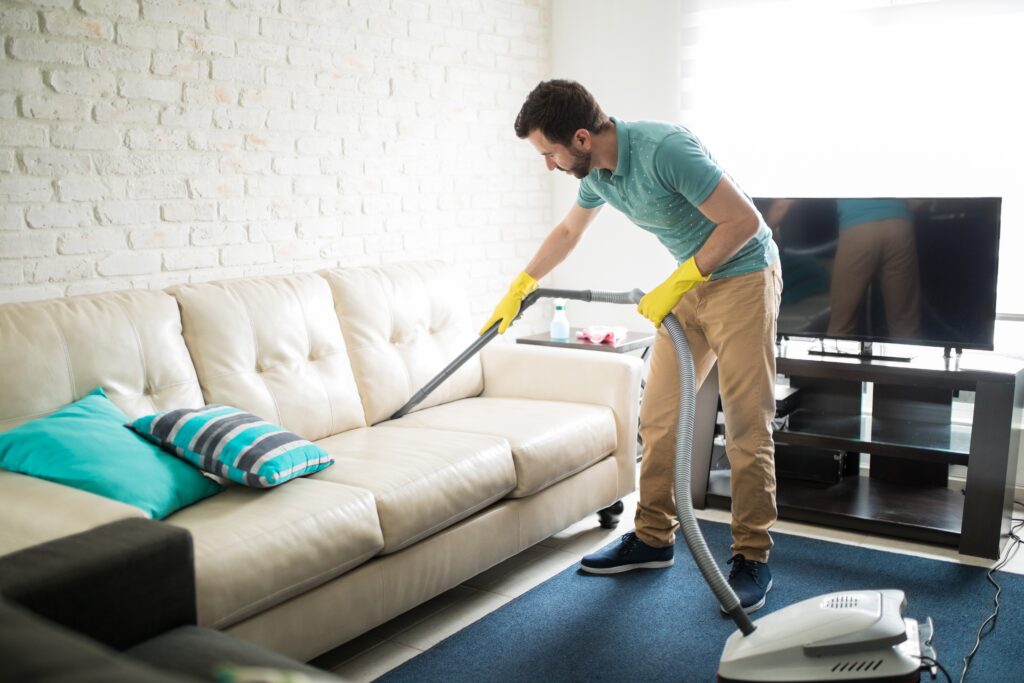 A young boy wearing green t shirt skin color trouser black shoes and yellow gloves standing on blue rug holding steam cleaner in hand and cleaning off white leather sofa 